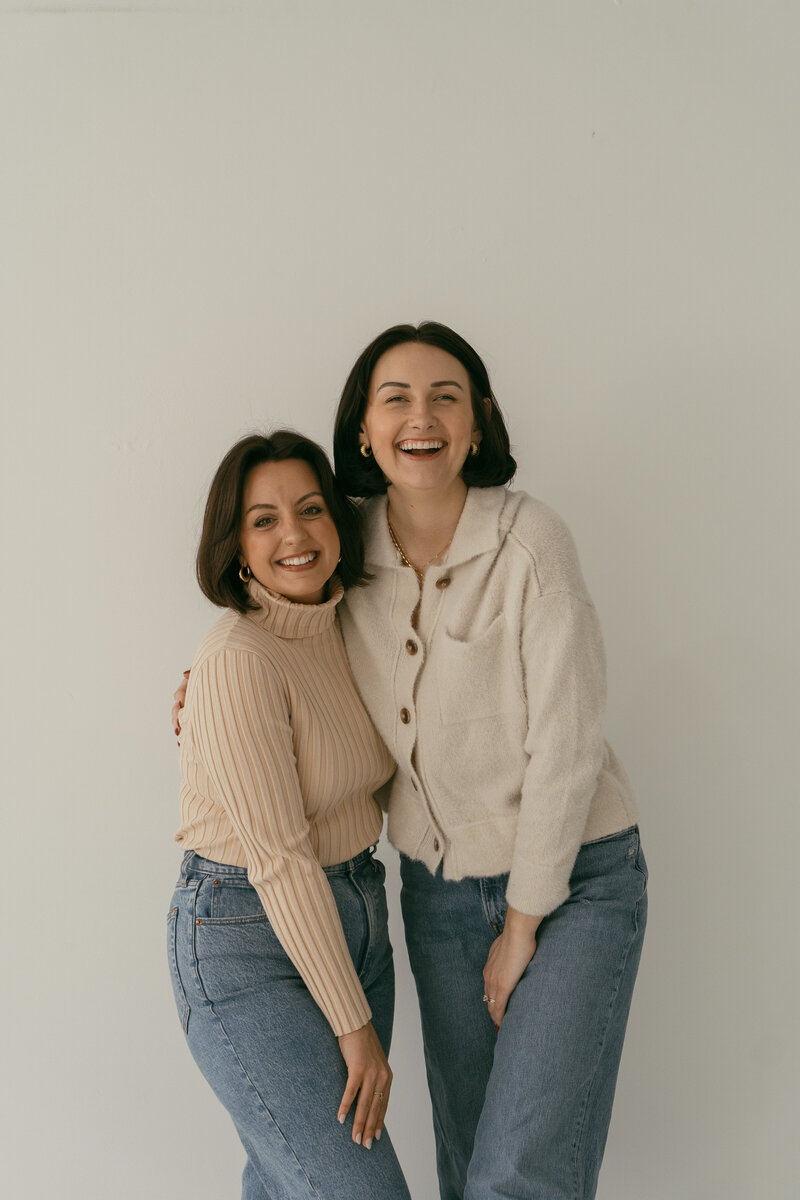 Two women smiling, posing together against a white wall.