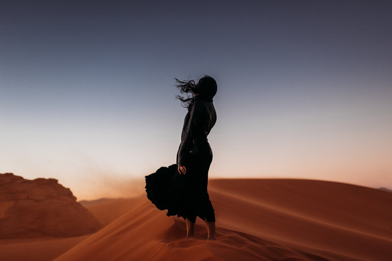 woman in black standing on a red sand dune with the wind blowing her dress and hair