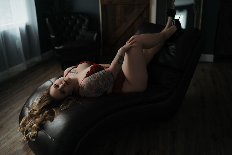 A woman wearing red lace lingerie lays across a chaise lounge with her black high heals in the air