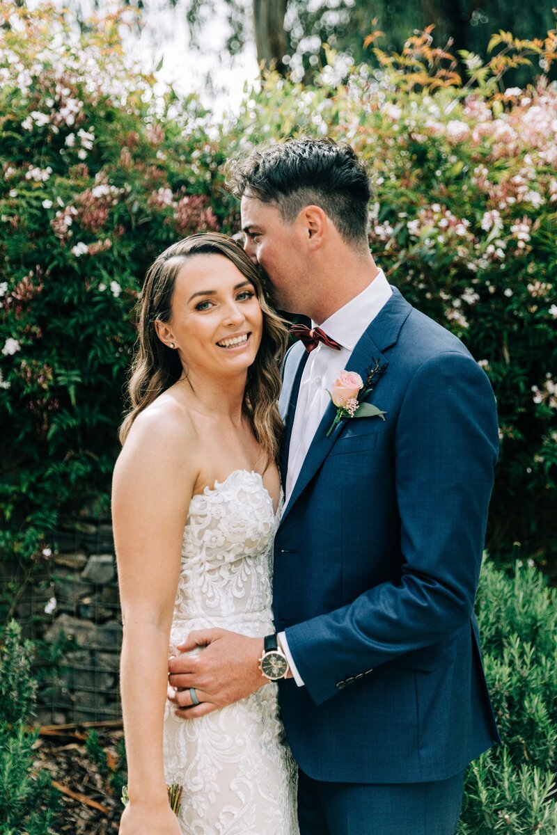 a bride wearing a strapless wedding gown staring in the camera and laughing while the groom holds her and his face partially hidden behind the bride as he whispers something in the bride's ears to make her laugh with a background of lush green gardens