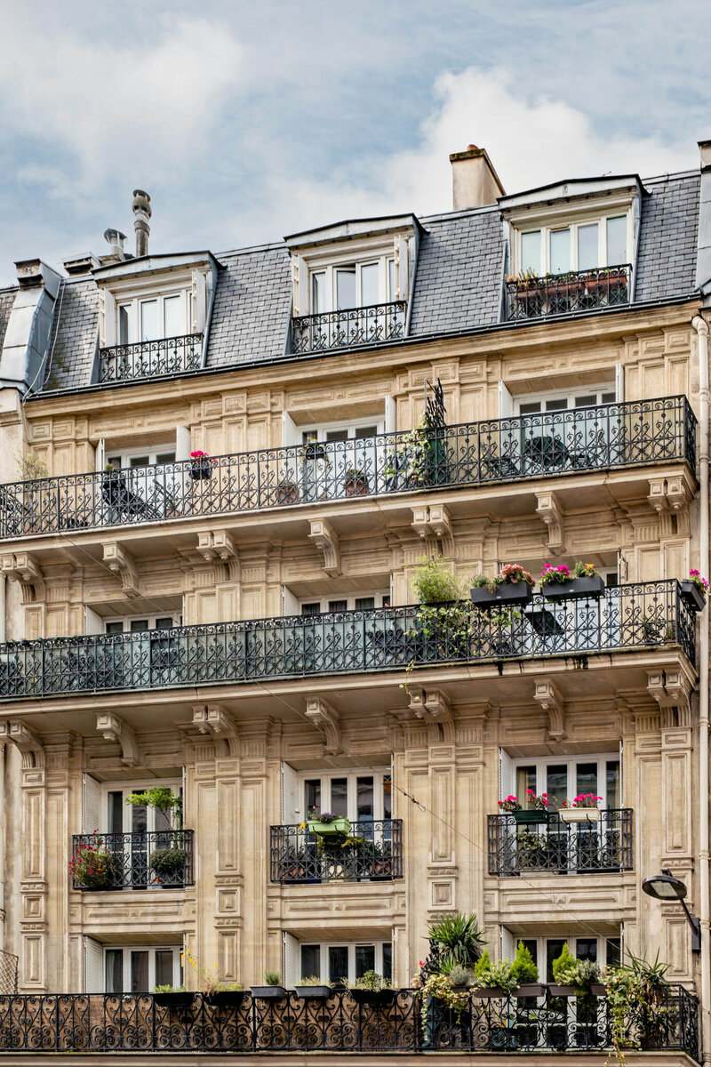 A classic European apartment building with ornate iron balconies adorned with potted plants. The facade features light stonework and a sloped slate roof. The balconies display various plants and flowers, adding color and greenery.