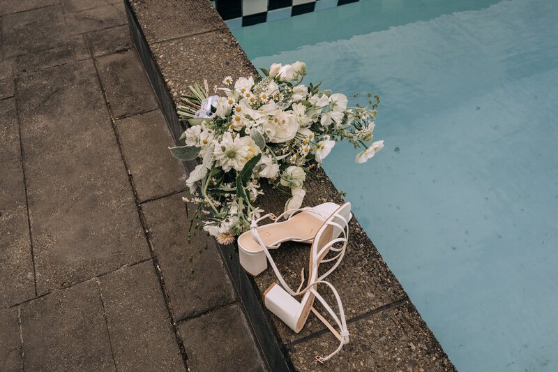 white sandal bridal heels and a green yellow and white unstructured wild bouquet lie beside a swimming pool in merivale