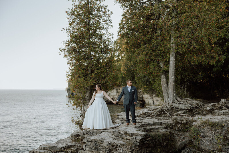 bride and groom standing on a rocky overlook in Door County with trees and Lake Michigan in the background.