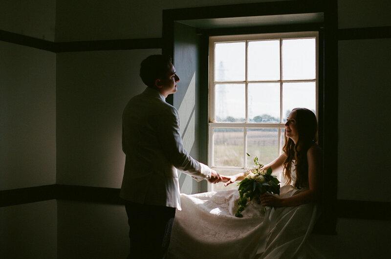 Bride sitting in windowsill holding grooms hand