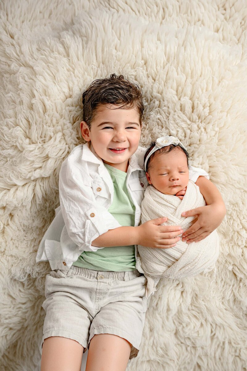newborn baby girl is snugged by her brother on a cream rug