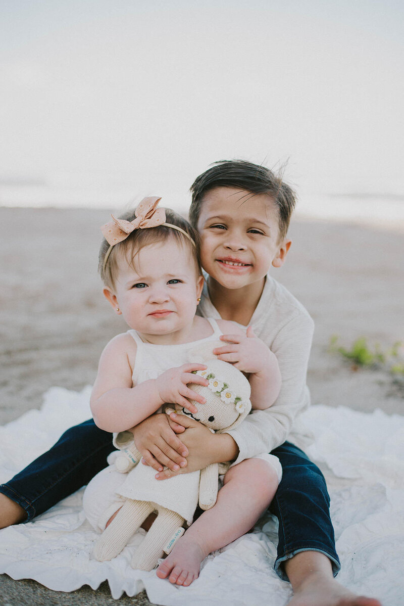 Toddler boy hugs his baby sister on the beach and smiles