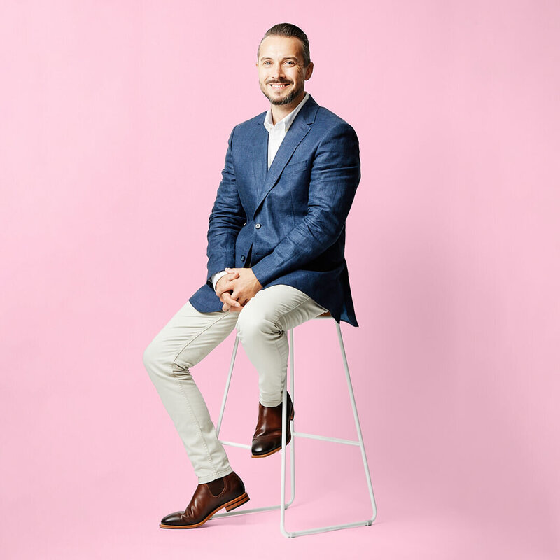 Man in a blue jacket on a white stool against a pink background.