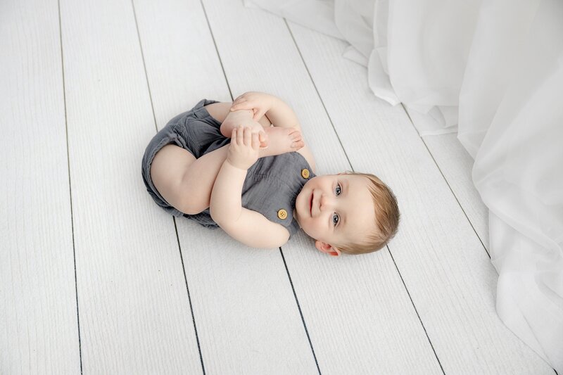 little boy looks toward camera durin ghis sitter session while smiling and playing with his feet