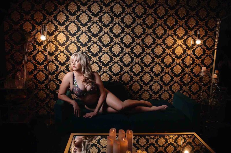 Older blonde haired woman posing on couch infront of black and gold baroque backdrop