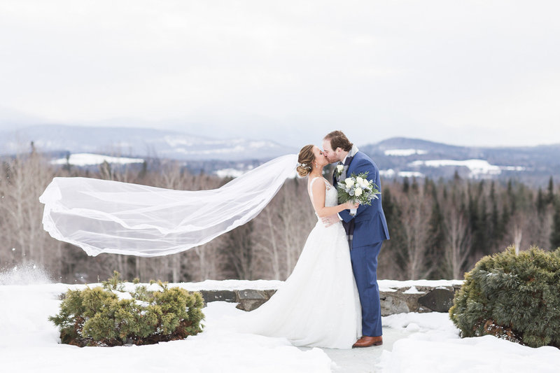 groom kissing wife with veil in wind on top of snowy mountain