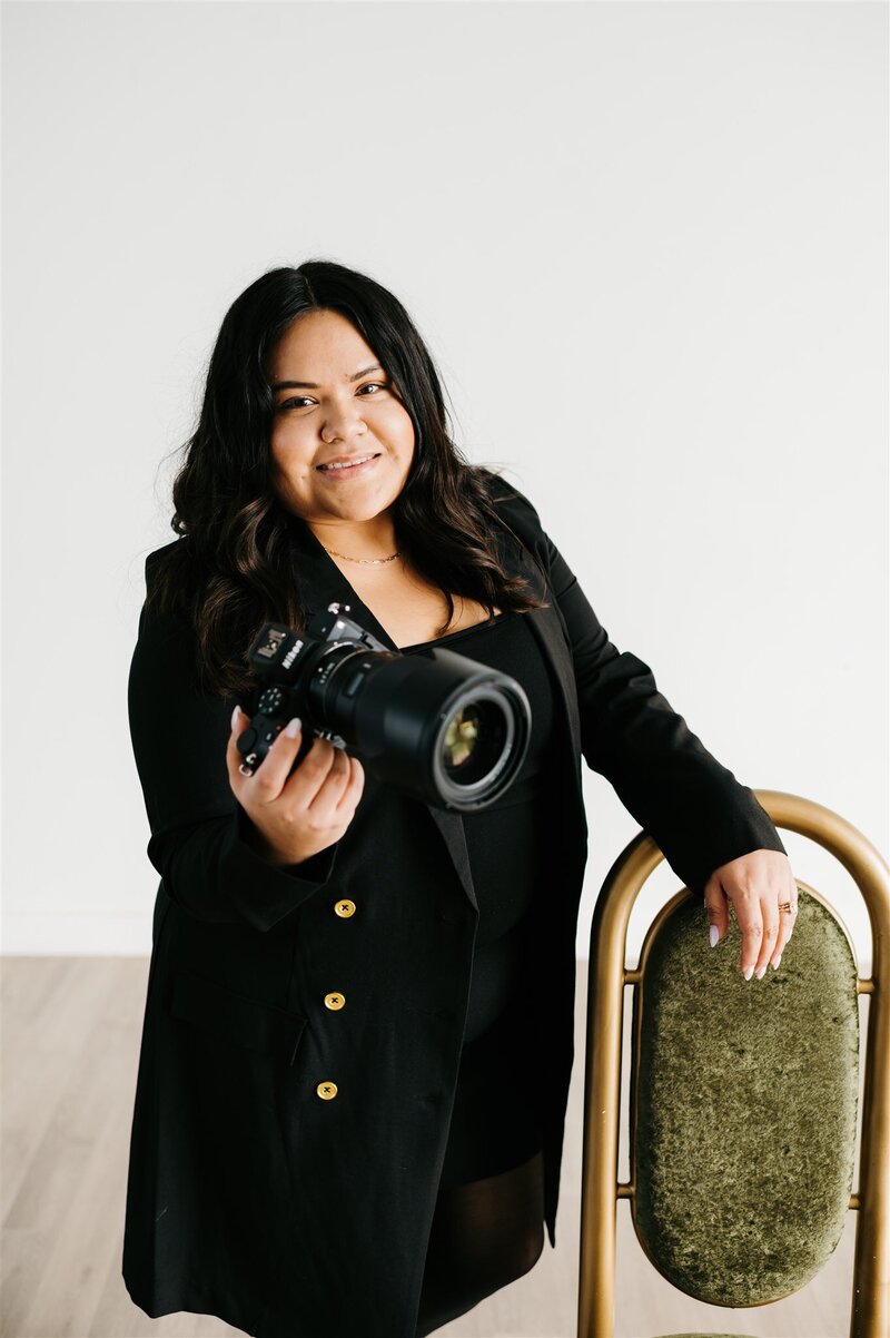 headshot of charlottesville wedding photographers  standing in a white studio and leaning one arm on a vintage green and gold chair while holding her camera in her hand