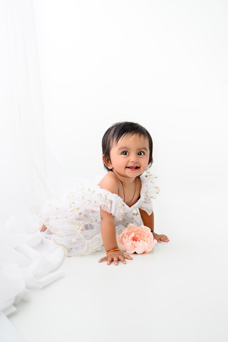 one year old girl crawling on her knees with a peach flower in front of her while wearing a white floral outfit