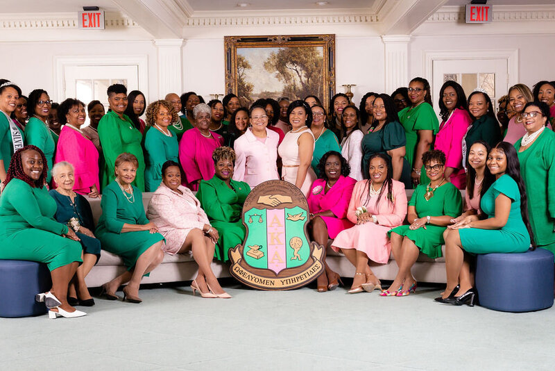 Phi Alpha Omega Chapter of Alpha Kappa Alpha wearing pink and green dresses gathered around the AKA shield in front of a landscape portrait