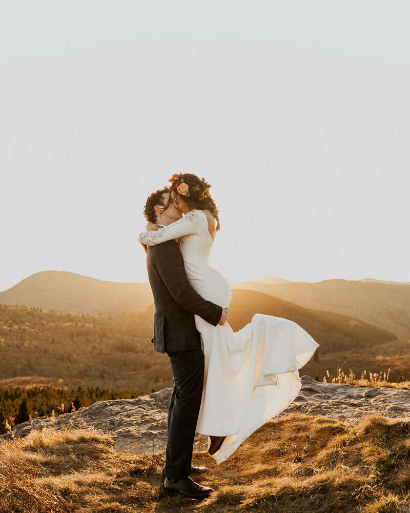 Groom in black suit lifts bride wearing pink flower crown up at golden hour in the mountains