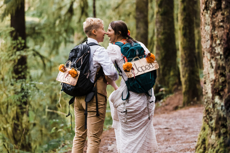 Two brides face each other laughing wearing backpacks with signs saying Just Married draped in marigolds as the rain pour down on their PNW adventure elopement day. | Erica Swantek Photography