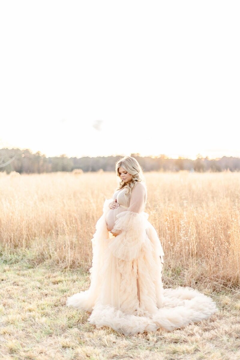 Woman in maternity gown stands in field of grass holding pregnant belly.