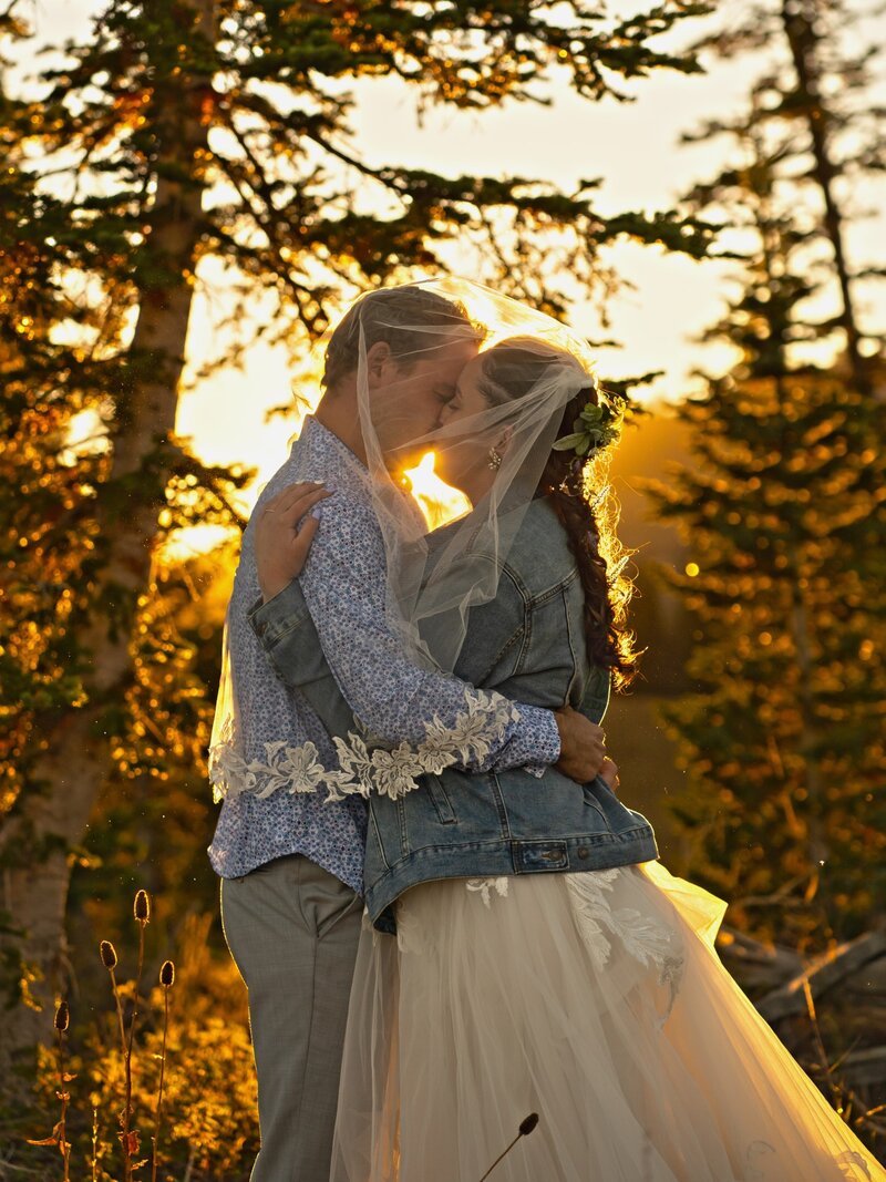 Enchanting photograph capturing a bride and groom sharing a heartfelt kiss amidst a forest backdrop, illuminated by the golden hues of sunset.
