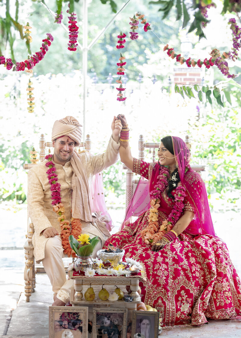 A couple in traditional South Asian wedding attire smiles and holds hands under a canopy adorned with hanging floral garlands. The bride wears a red and gold embroidered outfit, while the groom dons a cream-colored sherwani and turban.