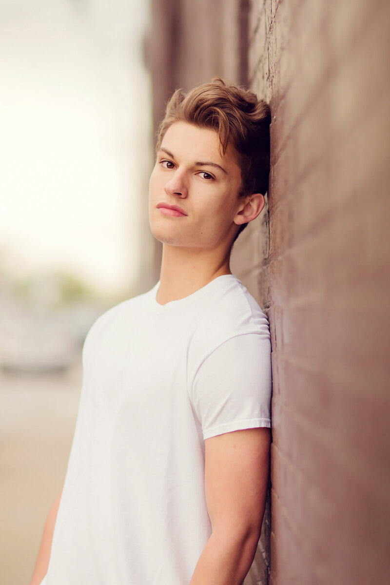 Senior portrait of a boy with brown hair and brown eyes leaning against a brick wall.