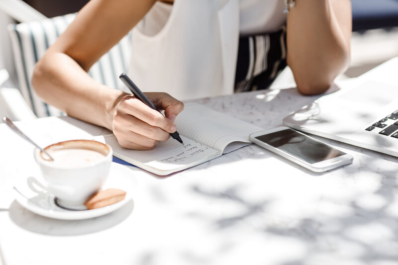 A person is writing in a notebook with a pen at an outdoor table, partially visible. Next to them is a coffee cup, a smartphone, and a laptop. The table is sunlit, with shadows of leaves creating patterns on the surface.