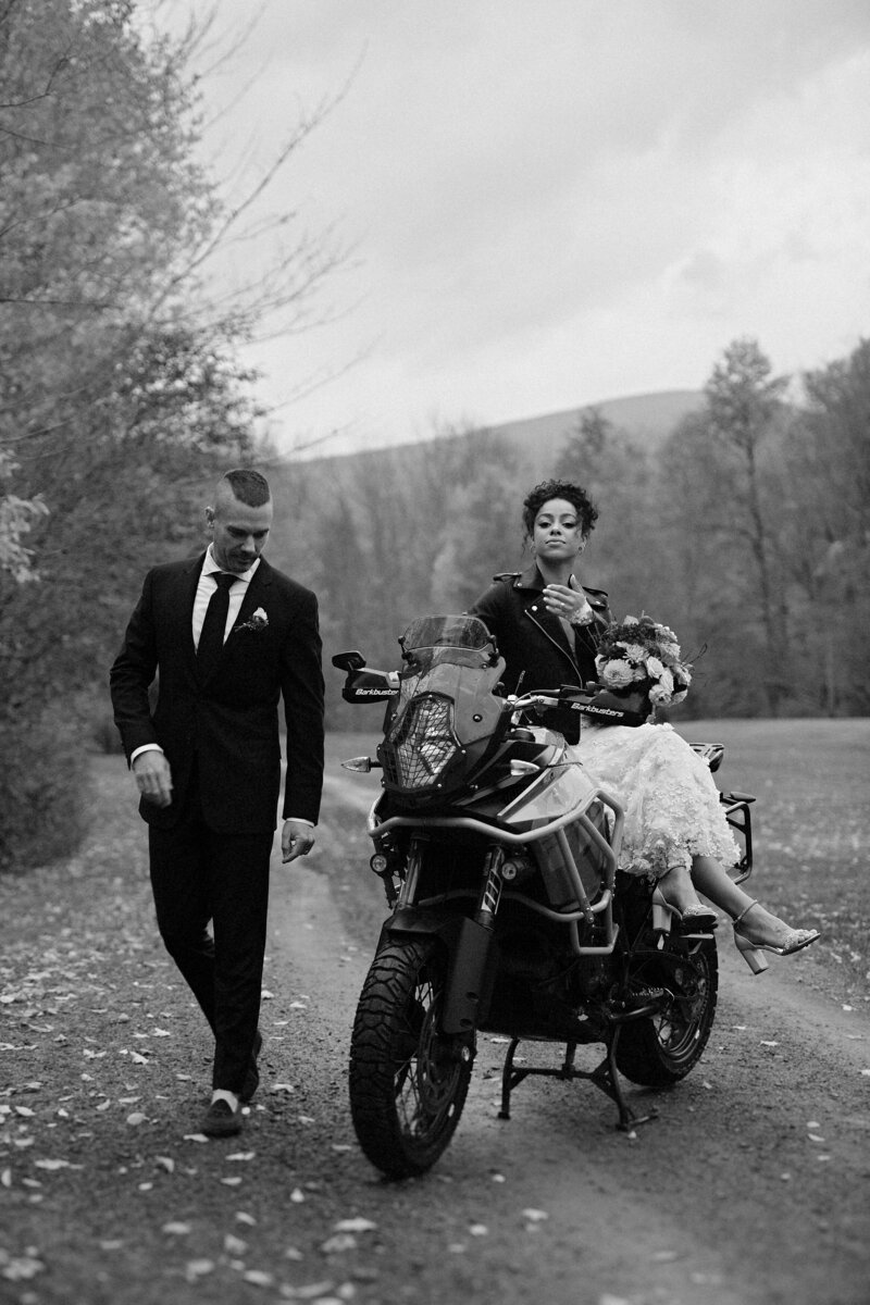 A groom walking alongside his motorcycle and a bride riding sidesaddle with her bouquet