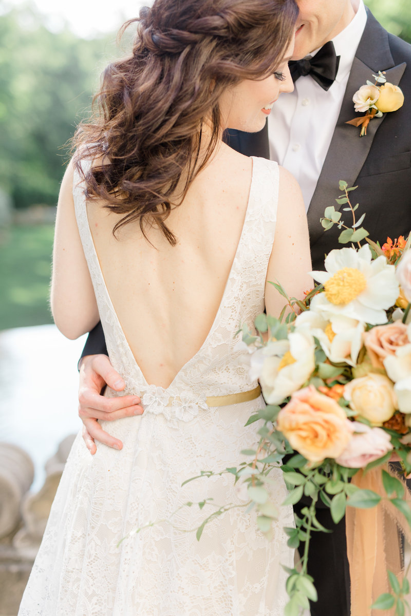 Bride and groom embrace and smile while holding full yellow and orange hued bouquet