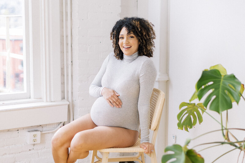 Photo of a pregnant woman wearing a gray bodysuit in a photo studio sitting on a chair next to a monstera plant, smiling at the camera