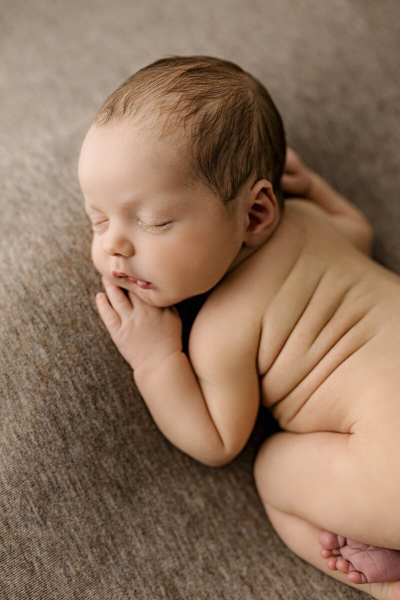Newborn baby boy overhead profile photo as he lays on a brown background where you can see his toes peeking out his little rolls of chub and his sweet lips and eyelashes and head full of hair