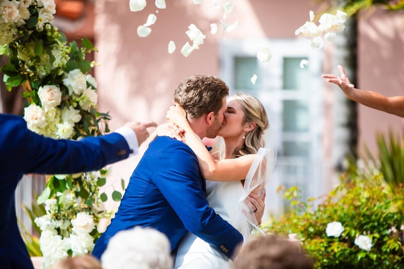 Bride and groom having their first kiss at their outdoor wedding ceremony in front of a floral arch with flower petals being thrown at La Valencia Hotel.