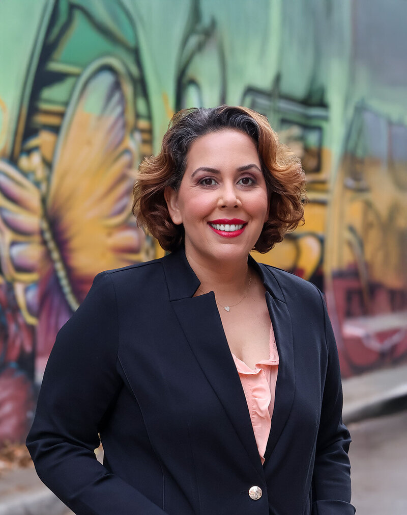 Ida Salusky with short, wavy hair in a dark blazer stands in front of a colorful graffiti mural featuring yellow and orange butterfly wings.