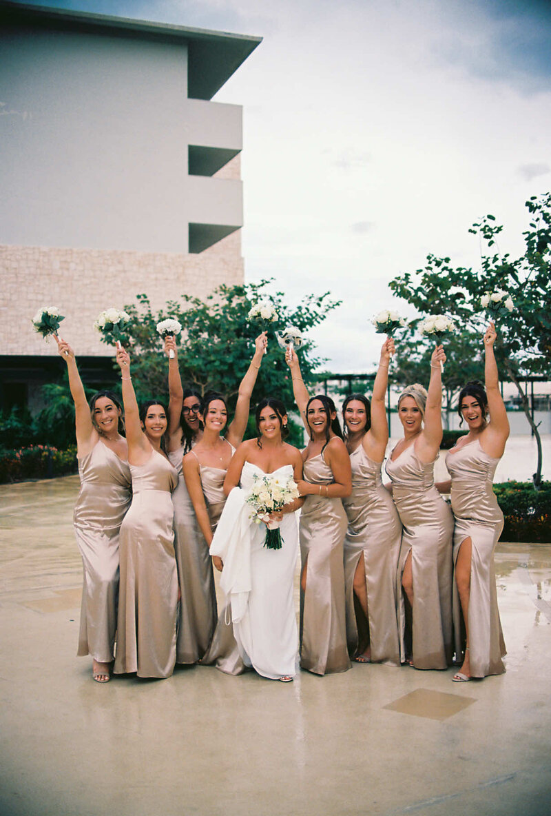 A bride in a white gown is surrounded by eight bridesmaids in matching champagne dresses, all holding bouquets high. The mood is joyful and celebratory.