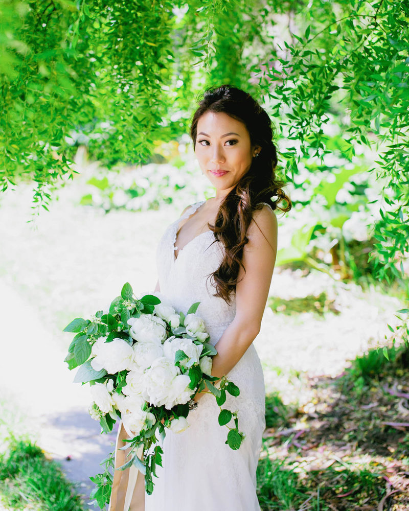 Bride holding bouquet standing under tree smiling at camera