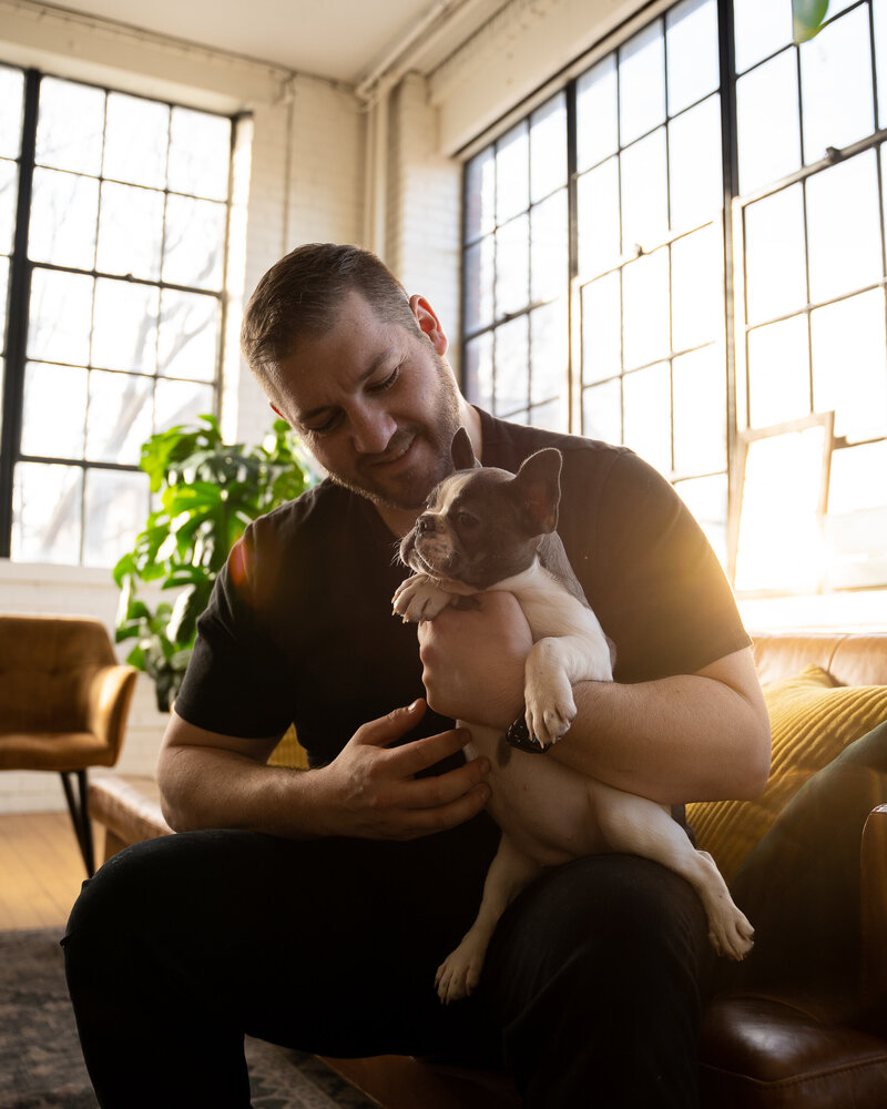 Frenchie Breeder Peter Kramm is sitting on a couch facing the camera holding a blue pied French bulldog puppy , He is holding the puppy looking down at her and smiling.