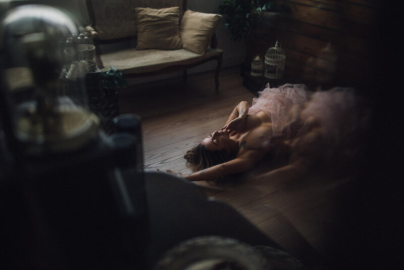 A woman lies on her back on the hardwood floor of a living room. Her top is bare and she wears a pink tutu. She emits light despite the room being dimly lit.