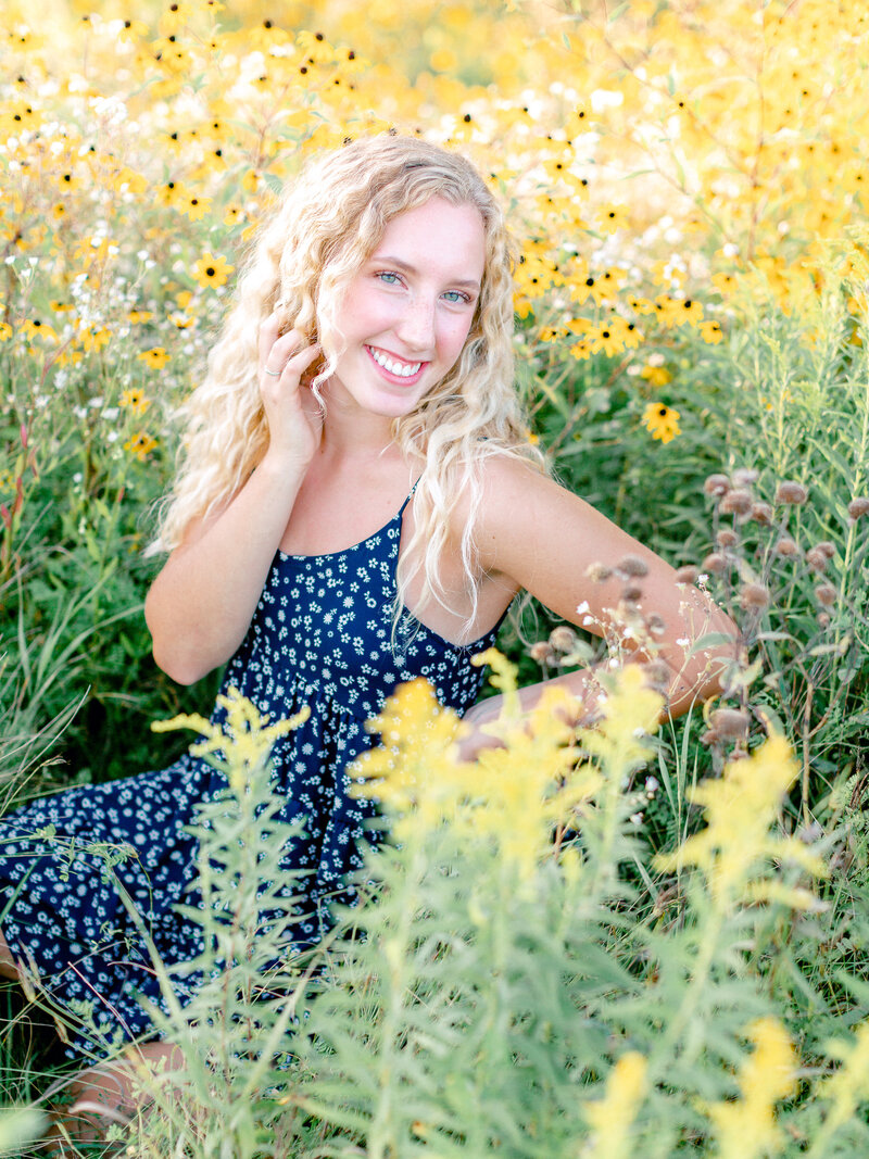 High school blonde senior smiling and sitting in a field of yellow wildflowers