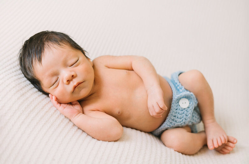 A newborn baby boy in a blue knit diaper cover sleeping on a white blanket during his natural light newborn session