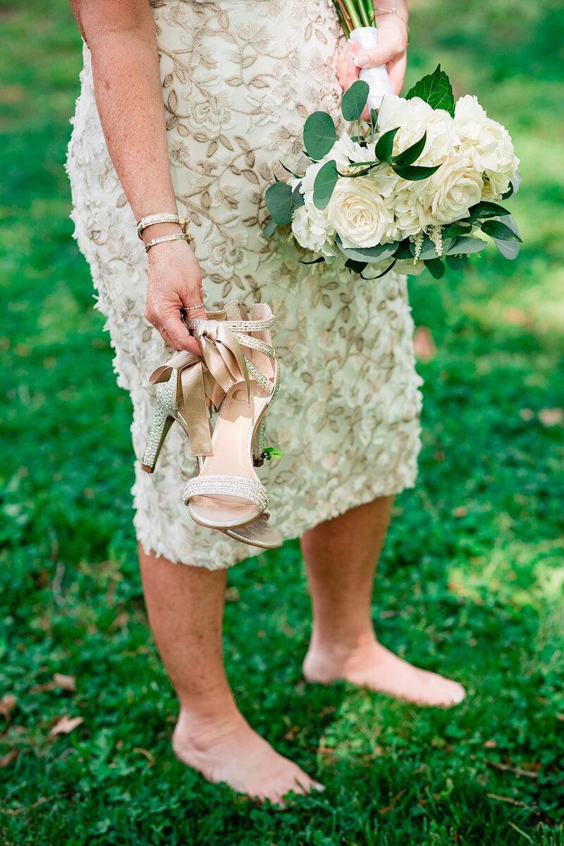 The bride goes barefoot in the grass removing her strappy stilettos. The bride is wearing a tea length lace dress and is holding a bouquet of white roses, white hydrangea and white peonies.