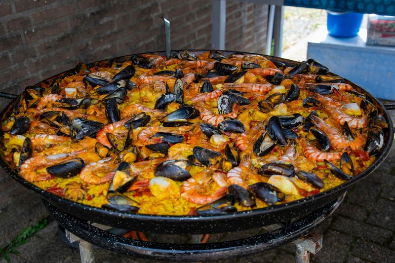 A large traditional Spanish paella in a wide, black pan cooking outdoors. The rice dish is filled with seafood including shrimp and mussels, displaying its characteristic yellow saffron color.