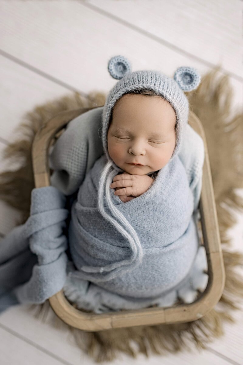 baby boy wrapped in blue with a bare Bonnet with little ears wrapped and gently placed in a crate on a wood floor