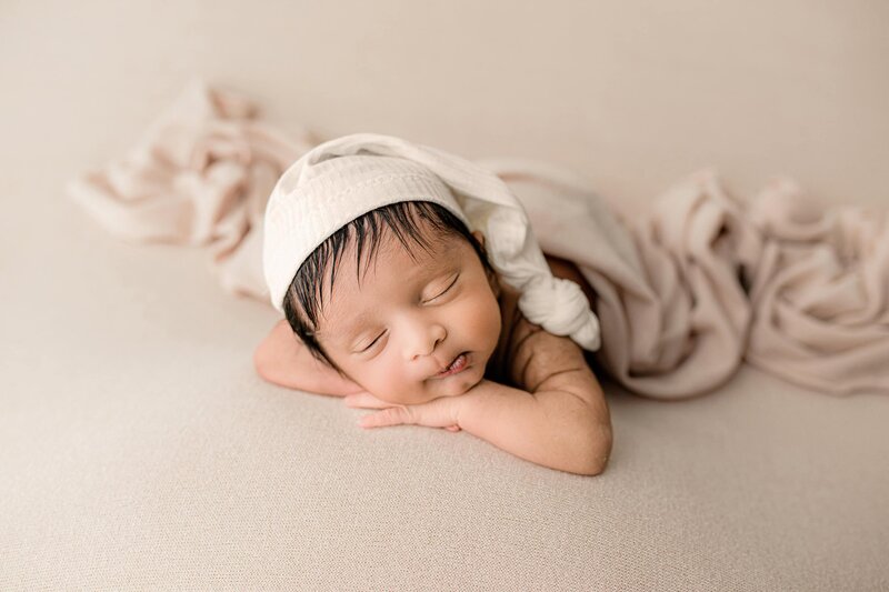 Newborn baby boy is wearing a sleepy cat placed on a neutral background with his hands both underneath his cheeks and elbows out to the side where you can see the close-up images and details of his face