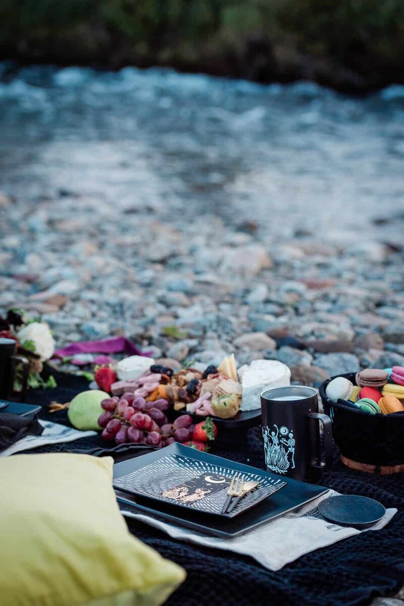 A beautiful styled picnic, arranged by a luxury destination elopement photographer is angled across the frame with all the colors of fruit, cheeses, and flowers.  There are brightly colored macaroons in a black bowl and black and white plates are set upon black blanket on a river rock beach, also of colorful stones. 