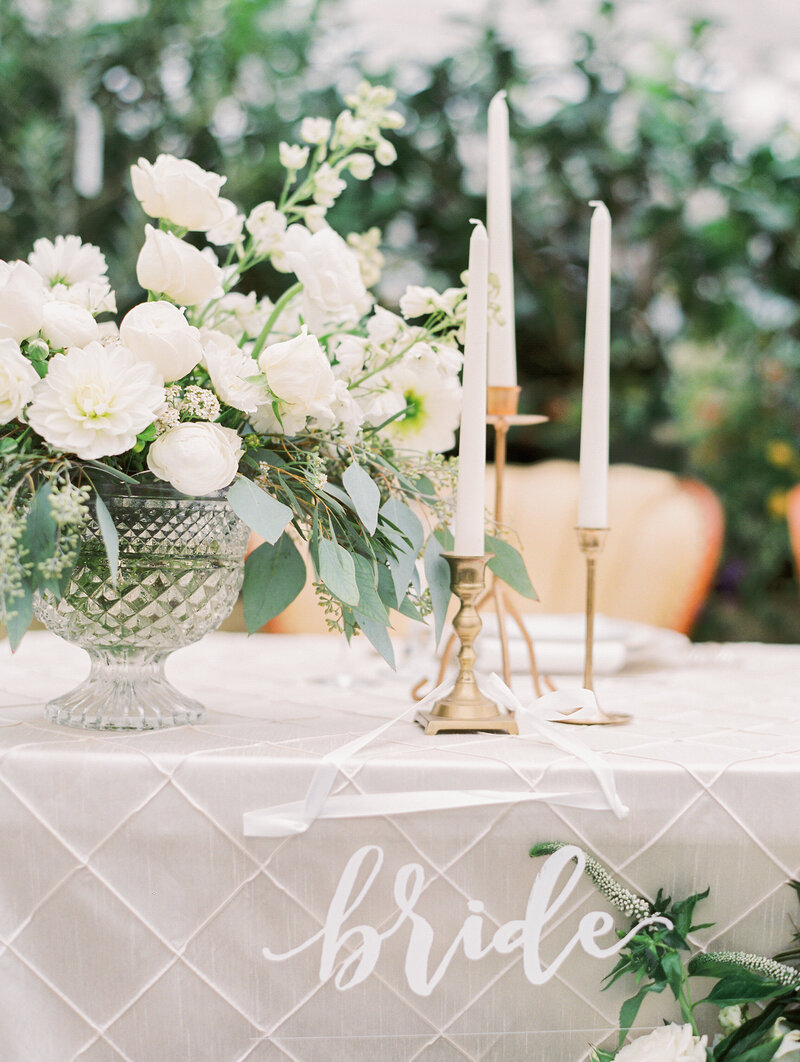 table with white candles and flowers with a bride sign on it