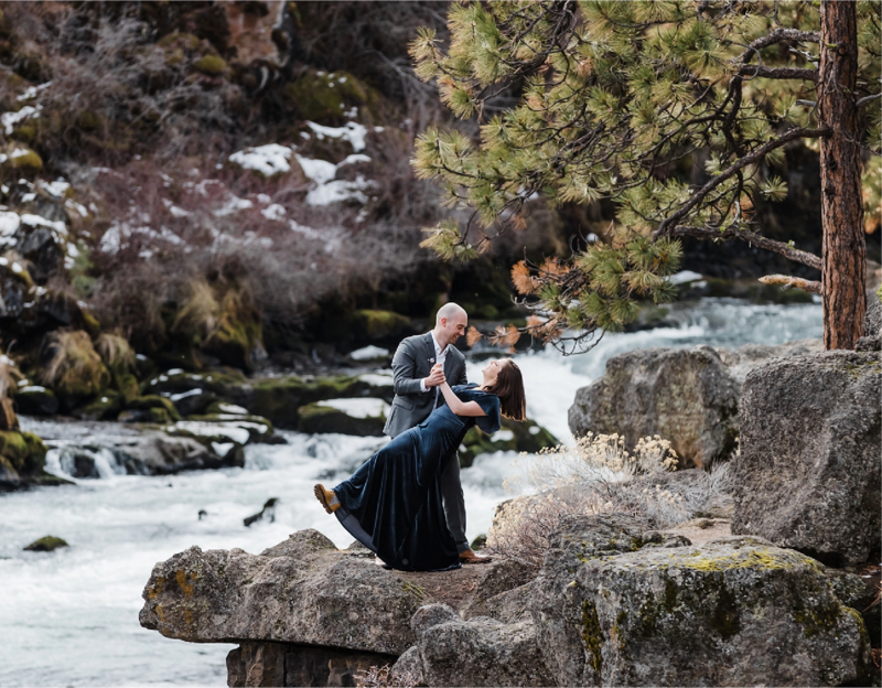 A bride wearing a blue velvet dress is dipped back while dancing with her groom on a rocky ledge perched high above Dillon Falls along the Deschutes River during their winter elopement in Bend, Oregon. | Erica Swantek Photography