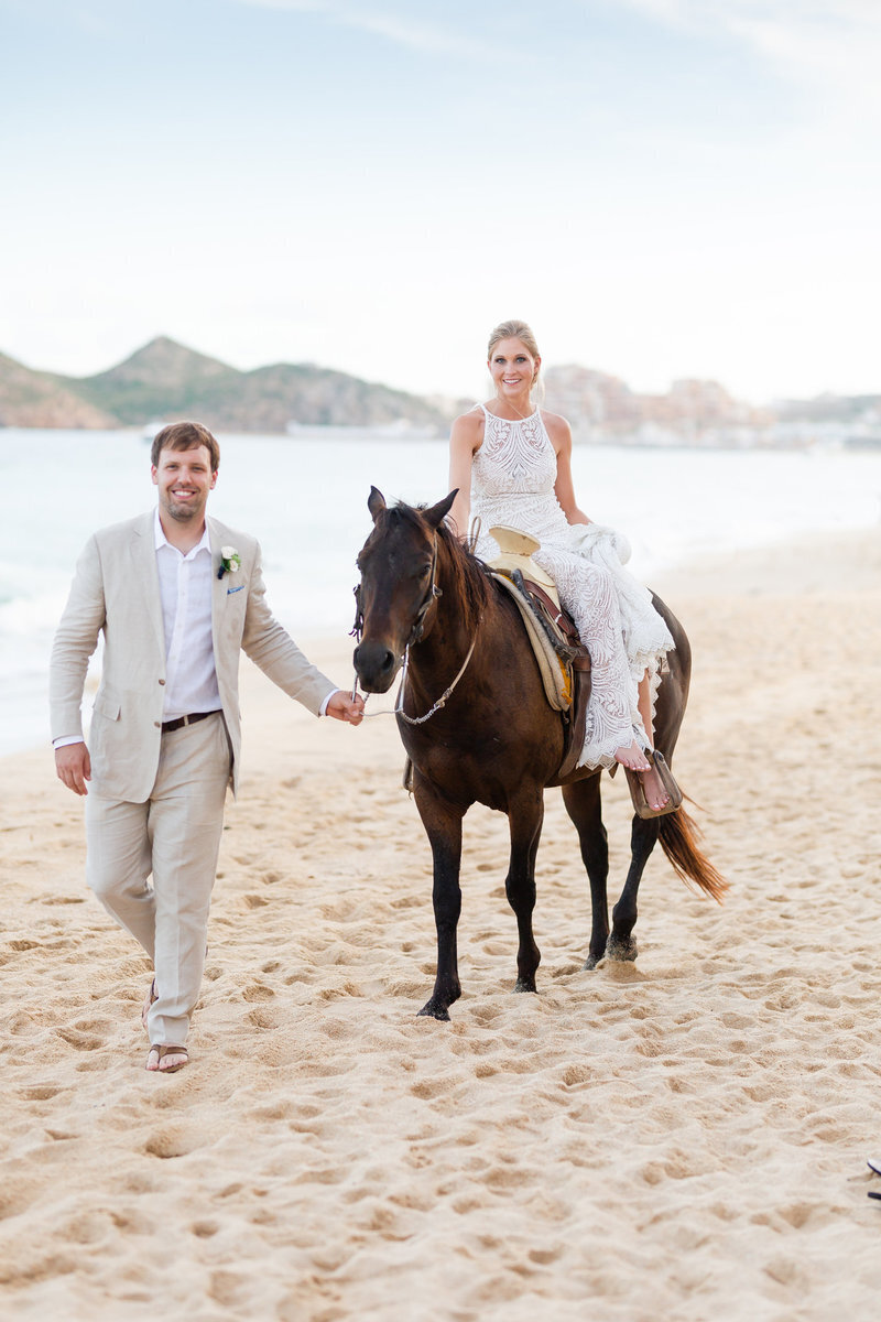 A bride riding a horse on the beach, accompanied by a smiling groom walking alongside, captured by a luxury wedding photographer.