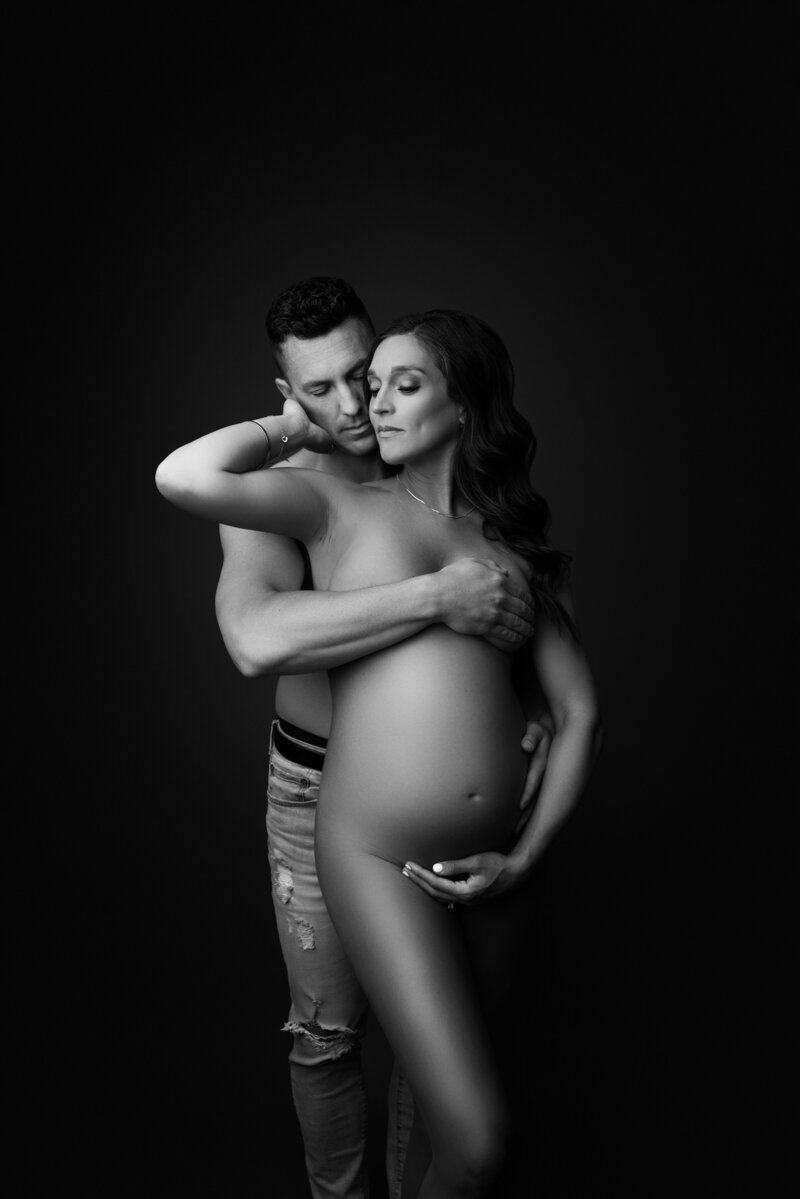 Black and white image of fine art Philadelphia maternity photoshoot.  Woman is standing bare facing the camera with her shirtless husband in jeans behind her. His arm is covering her breasts and the other hand on her hip. The woman's hand is under her bump and the other arm is reaching back and touching her partner's cheek. The woman is turning her face toward her partner. Their faces are touching. Captured by best Philadelphia and Main Line maternity photographer Katie Marshall.