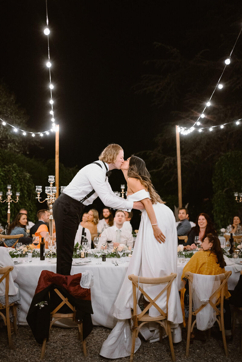 Bride and groom kiss standing on chairs during a wedding dinner