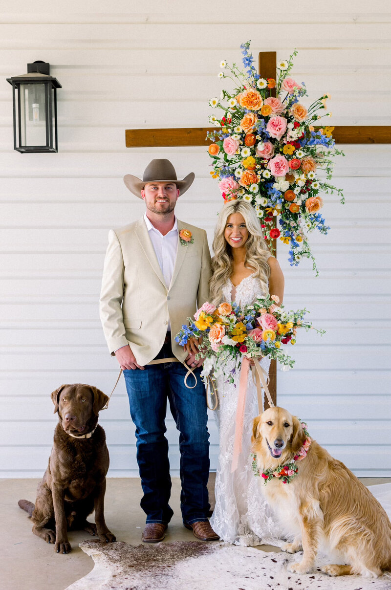 a colorful flower arrangement on a wooden cross with two dogs sitting on a cowhide rug at wedding ceremony
