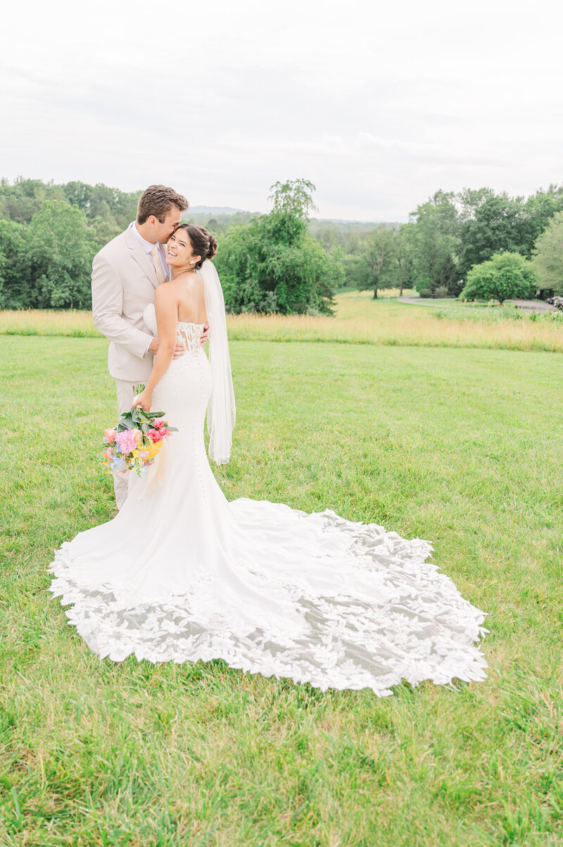 A laughing brunette bride with her husband as he kisses her cheek in a bright green field in Jun by JoLynn Photography