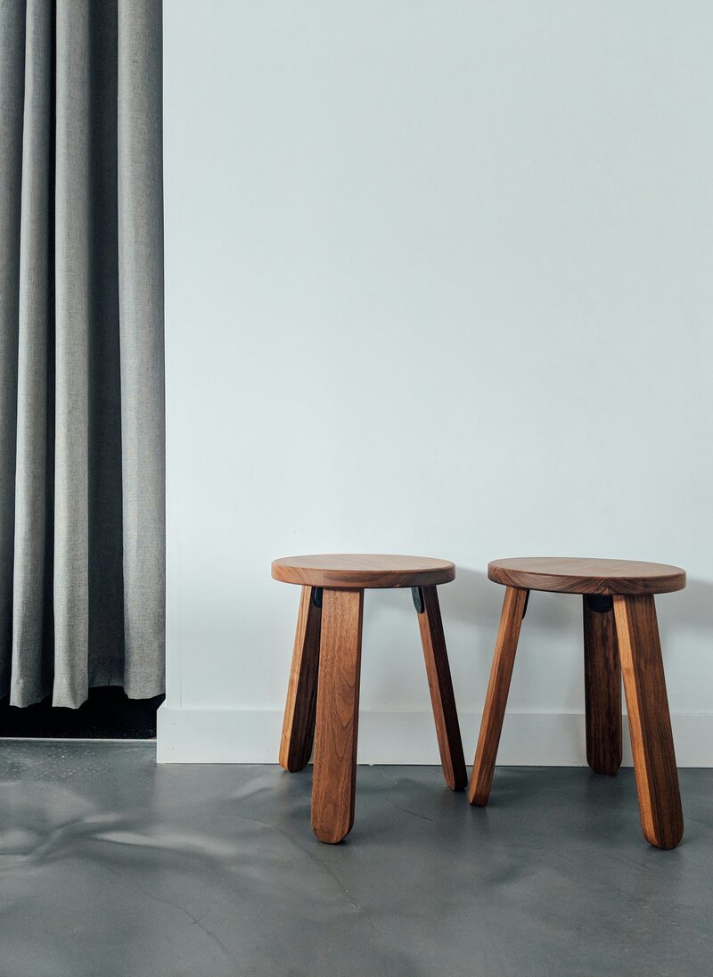 Two wooded stools sitting together in front of white wall with dark grey concrete floor in design studio