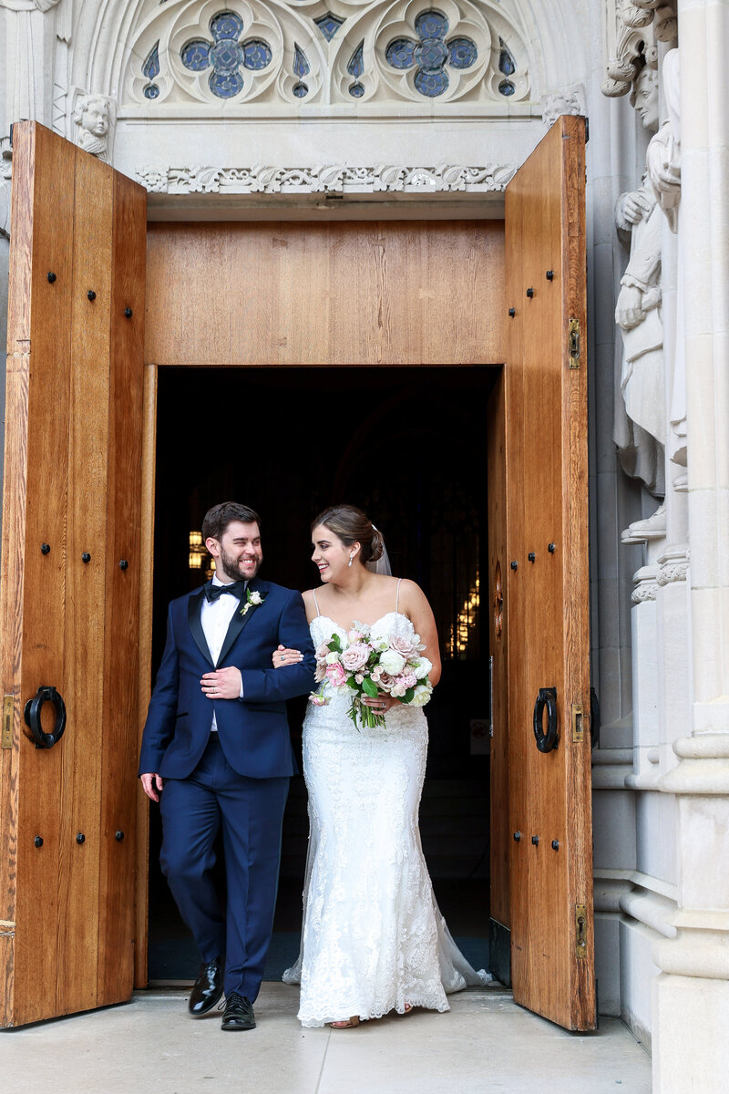 Bride in a spagetti strapped dress with lace detailing and assorted pale pink flowers holding arms of blue navy tux and bowtie  walking out of duke chapel  in durham nc